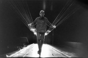 A series of strings are stretched across a room. A person stands between them and plucks them.