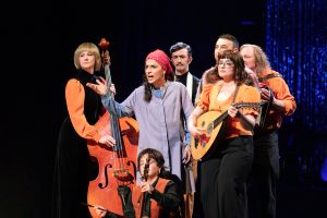 A woman wearing a headscarf and a work coat stands in the middle of a group of musicians and speaks to the audience.