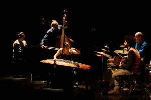 Fünf Musiker*innen mit verschiedenen Instrumenten auf einer dunklen Bühne.