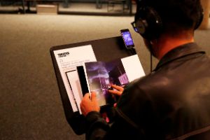 Over the shoulder of a person wearing a dark shirt and glasses, hands can be seen leafing through a book of photos.