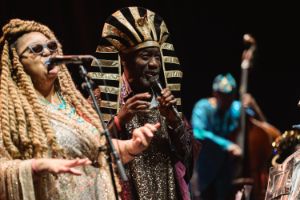 Das Sun Ra Arkestra auf der Großen Bühne im Haus der Berliner Festspiele: im Vordergrund Tara Middleton singend am Mikrofon, rechts daneben Michael Ray mit einem schwarz-goldenen Kopfschmuck.