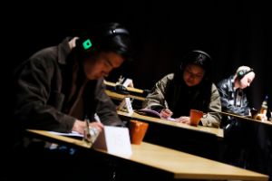Three people wearing headphones are standing at high tables and writing something on pieces of paper.