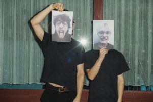 Zwei Personen in schwarzer Kleidung stehen nebeneinander und halten jeweils ein ausgedrucktes Foto mit einem Porträt vor ihr Gesicht.