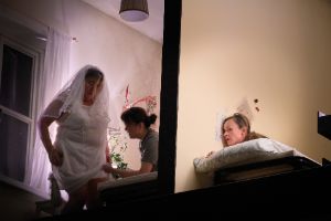 A woman listens at a bright yellow wall behind which a bride is being dressed.