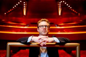 A man sits at the conductor’s podium in a large concert hall and looks past the camera into the room.