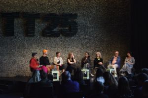 Eight people are sitting on a podium talking to each other, with the audience in the foreground.