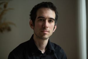 A man with short black hair and a black shirt looks kindly into the camera.