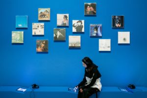 Eine Person mit Kopfhörern sitzt auf einer Bank vor einer blauen Wand, an der Schallplatten-Cover hängen.