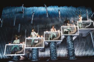 Five people perform on five rising Plexiglas platforms, surrounded by smooth tubes and ribbed pipes.