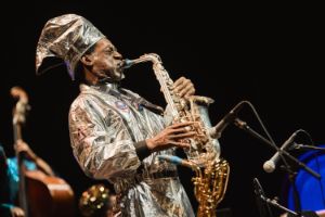 Knoel Scott im Profil, er spielt Saxophon, trägt einen goldenen Kopfschmuck und einen goldenen Overall.