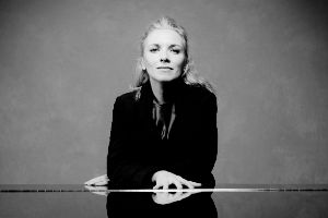 Eine Frau mit blonden Haaren in einem schwarzen Samtblazer steht hinter einem zugeklappten Klavierflügel und schaut in die Kamera.