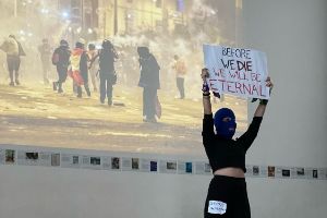 Eine vermummte Person in schwarzer Kleidung steht vor einem Foto eines Platzes, auf dem eine gewaltsame Auseinandersetzung stattfindet, und hält ein Schild mit dem Text „Before we die we will be eternal“ hoch.