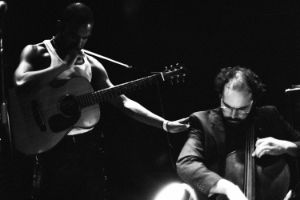 Schwarz-Weiß-Foto von zwei Personen mit Instrumenten auf der Bühne. Die eine Person hat  eine Gitarre umgebunden, die andere spielt sitzend Bass.