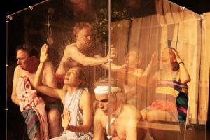 Das Ensemble sitzt in einem Glaskubus und ist in Handtücher gekleidet. Die Situation erinnert an eine Sauna.