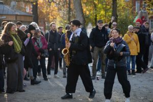 Zwei Musiker*innen mit Saxofon und Trommel gehen an einer Publikumstraube vorbei. Sie befinden sich im Freien.