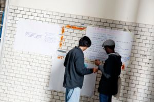Zwei Personen stehen vor einer Wand, an der große Zettel mit handschriftlichen Notizen hängen.