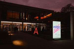 Außenansicht des Haus der Berliner Festspiele am Abend, vor dem Haus leuchtet ein rotes A und das Plakat des Jazzfest Berlin 2024.