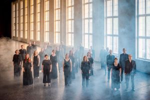 The Helsinki Chamber Choir stands in a large, industrial hall. Blue and yellow light is coming through the large windows, which becomes visible in the light fog surrounding the singers.