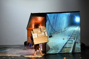 Ein Mensch mit nackten Beinen trägt Kartons vor einem Haus, an das Videomaterial von einem Tunnel projiziert wird.