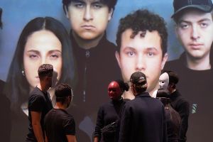 Schüler*innen spielen Theater, haben Masken auf, stehen im Kreis. Im Hintergrund eine Leinwand mit ihren unverdeckten Gesichtern.