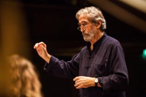 A man with grey hair and glasses is conducting, his right hand raised.