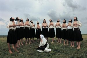 Women wearing black headscarves and knee-length skirts massage their bare breasts. As they do so, they gaze up at the sky.