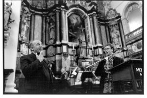 An orchestra is playing in a church. Two men are standing in the foreground.