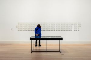 Eine Person steht in einem Ausstellungsraum vor vielen kleinen Rahmen, darin sind kleine Zettel zu erkennen.