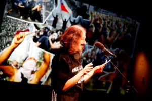 A person with shoulder-length red hair and a fake black mustache stands at the microphone and reads a text. Images of protest movements are projected in the background.