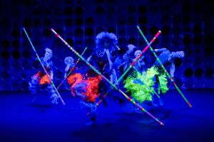 Die rüschigen Kostüme und langen Stäbe von den Tänzer*innen leuchten in bunten Neonfarben.