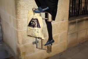 The image shows a two-tiered metal frame mounted into a stone wall. The lower half of a person wearing a black skirt and blue shoes is visible, stepping onto the frame. Hanging from it is a light yellow plastic bag with Hebrew lettering.