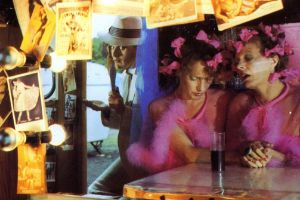 A film still showing a mirrored reflection of two women-presenting figures dressed in pink with ribbons in their hair, seated at a table. A man-presenting figure wearing a white hat stands in the doorway, watching them.