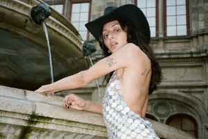 A close-up of a woman in front of a building with a black cowboy hat and silver dress looking into the camera.