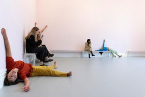 In einem hellrosa gestrichenen Raum findet eine Live-Performance mit dem Titel „study now steady“ statt. Mehrere Tänzer:innen bewegen sich dynamisch durch den Raum und beziehen dabei den Boden, die Wände und die Bänke in ihre Choreografie ein. Das Publikum sitzt auf einfachen Bänken und beobachtet aufmerksam das Geschehen.