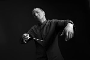 Ein Mann mit schwarzem Hemd steht dirigierend mit einem Taktstock in der Hand und geschlossenen Augen vor der Kamera.