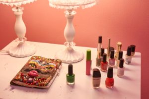 The picture shows a white nightstand in front of a pink wall with two lamps, a notebook and several bottles of nail polish on it.