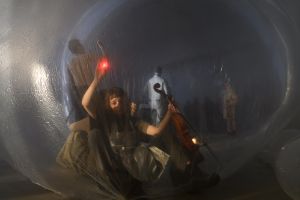 A woman holding a red lamp in her right hand and a violin in her left sits in a large transparent sphere. Other people stand in the background.