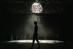 Eine Person steht im Gegenlicht vorne auf der Bühne, im Hintergrund mehrere Personen, die um einen Lichtkegel herum stehen.
