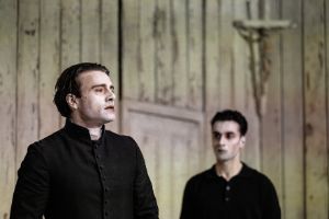 Two serious-looking men in front of a wooden wall on which hangs a small cross with Jesus.