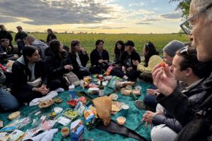 A group of young people are sitting on blankets on a lawn, eating and chatting.