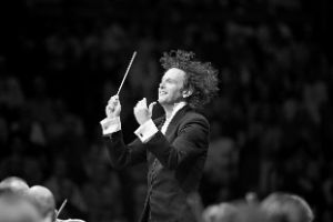 A man with curly hair and a baton in his hand conducts so exuberantly that his hair swirls around.