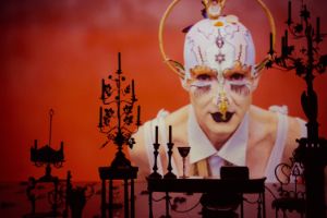 A projection of Cohen with delicately done make-up; in the front there are orante candelabrums