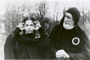 A girl forms a picture frame in front of her eyes with the thumbs and index fingers of both hands. An older man standing next to her looks at her. Both are wearing black cloaks.