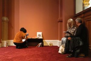 In der Ecke eines Raumes mit Teppichboden aus dunkelrotem Samt, befindet sich ein Bereich mit Büchern. In dieser Bücherecke stöbern Besucher*innen der Ausstellung "Ligia Lewis: I’M NOT HERE FORRRRR…" in zur Ausstellung thematisch passender Literatur.