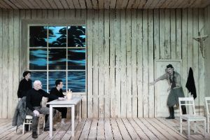 Three people are sitting in a room made entirely of wood, looking towards a person with an arm sling on the door.