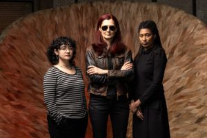 Das Trio steht in aufrechter, taffer Pose vor einem Holzobjekt.