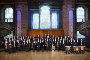 Musiker*innen eines Orchesters stehen mit ihren Instrumenten in einem kirchenähnlichen Gebäude.