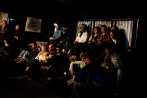 Audience members sit in a small auditorium and laugh about what they see. A drawing of a monster's head hangs on a black wall at the side.