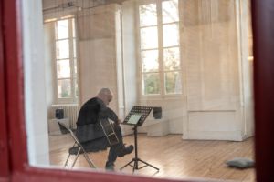 Ein Mann mit Gitarre sitzt in einem Raum mit vielen Fenstern im Sonnenlicht.