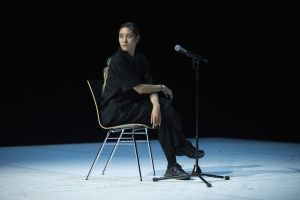 Das Foto zeigt eine weiblich gelesene Person, die mit überschlagenen Beinen auf einer Bühne auf einem Stuhl sitzt und ein Mikrofon vor sich stehen hat. Der Hintergrund ist schwarz.